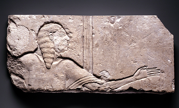 Limestone relief carving from ancient Egypt of an aged courtier (ca. 1336-1352 BCE). Fragment of a figure's face in profile, chest, and extended left arm showing thin form, aging skin, and frail hand.