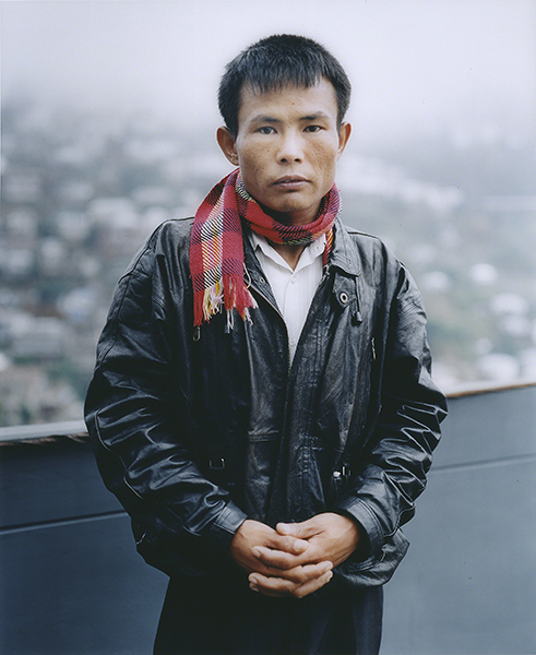 Photograph by Chan Chao titled Soy Myint (1997). Portrait of a man wearing a black leather jacket and red plaid scarf in front of a soft-focused hillside.