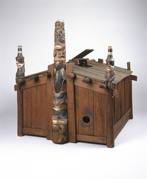 Carving by George Dickson titled Model of House of Contentment (1890s). Wood model of a Haida clan house with totem pole in the center of the facade and corner posts with watchmen figures.
