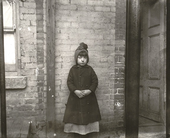 Photograph by Jacob Riis titled "I Scrubs" - Katie (1890). Black-and-white photograph of a young girl wearing a dress, coat, and hat and standing in front of a brick wall.