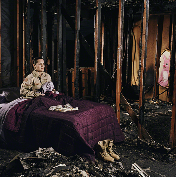 Photograph by Jennifer Karady from the series In Country: Soldiers’ Stories from Iraq and Afghanistan (2010). Woman in military uniform sits up in a bed with a pile of money on it in a room destroyed by an explosion with a pink bunny costume visible on the right.