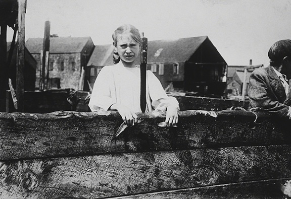 Photograph by Lewis Wickes Hine titled Minnie Thomas (1911). Photograph of a young girl standing behind a low wood wall with a large knife for cutting sardines.