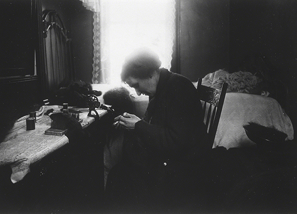 Photograph by Lewis Wickes Hines titled Tenement Home Worker Making Hairpieces at Home (ca. 1910). Black-and-white photograph of a woman sewing in front of a window.