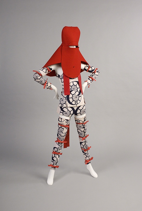 Outfit by Yamamoto Kansai titled Woman's Playsuit (1971). Mannequin wearing red felt hood with eye slit covering the face and shoulders and shorts, halter top, arm guards, and leg guards in white fabric with blue printed design and red ribbons.