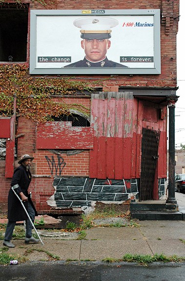Photography by Zoe Strauss titled Marines Billboard, Philadelphia (2004). Man with crutches walks by boarded up building with a billboard of a photo of marine and the text "the change is forever."