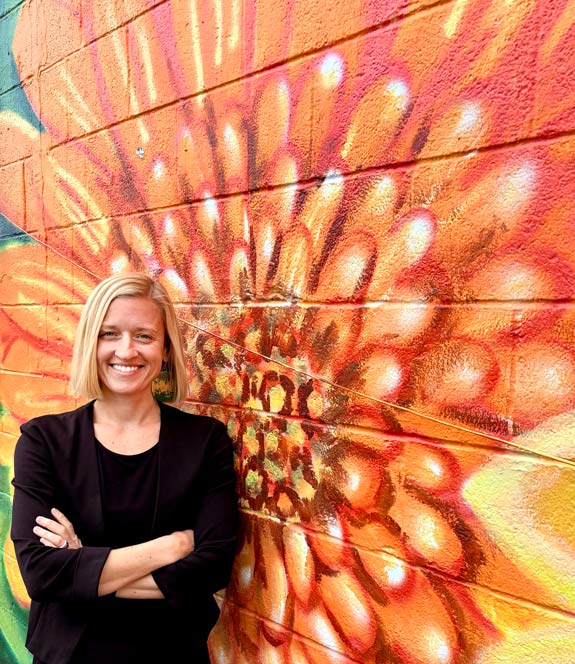 Dr. Leigh Drake in front of a vibrant flower mural by Tamara Vogrin Tara in Virginia Beach’s ViBe Creative District.