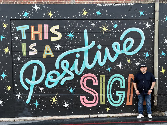 Frank stands in front of the mural This Is a Positive Sign by Peter Parpan in Asheville, North Carolina. The mural was funded by The Booth Fairy Project. Photo by Mary Lou Mayr.